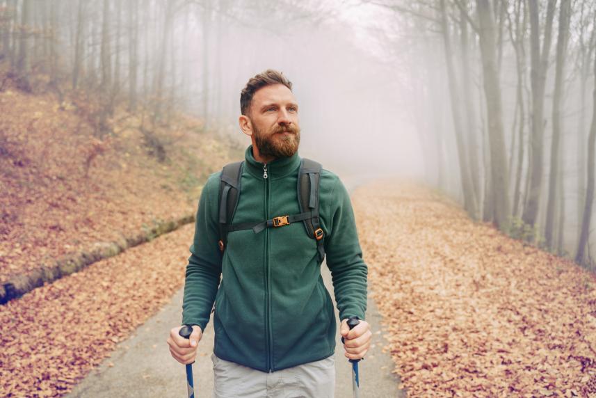 A blue-eyed, brunette man hikes up a leaf-covered road, using two walking sticks. He is dressed in a forest-green fleece and wearing a backpack.